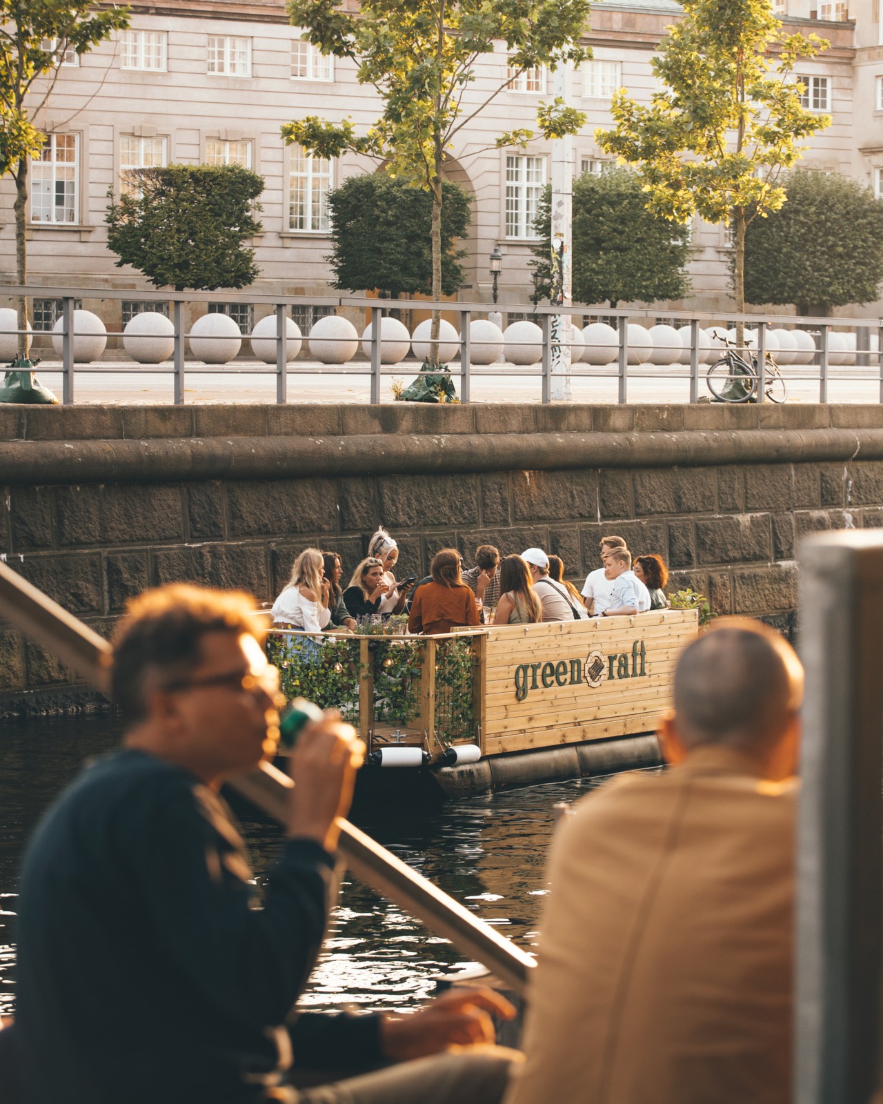 En Green Raft-tømmerflåde sejler forbi, mens folk sidder på kajen og kigger