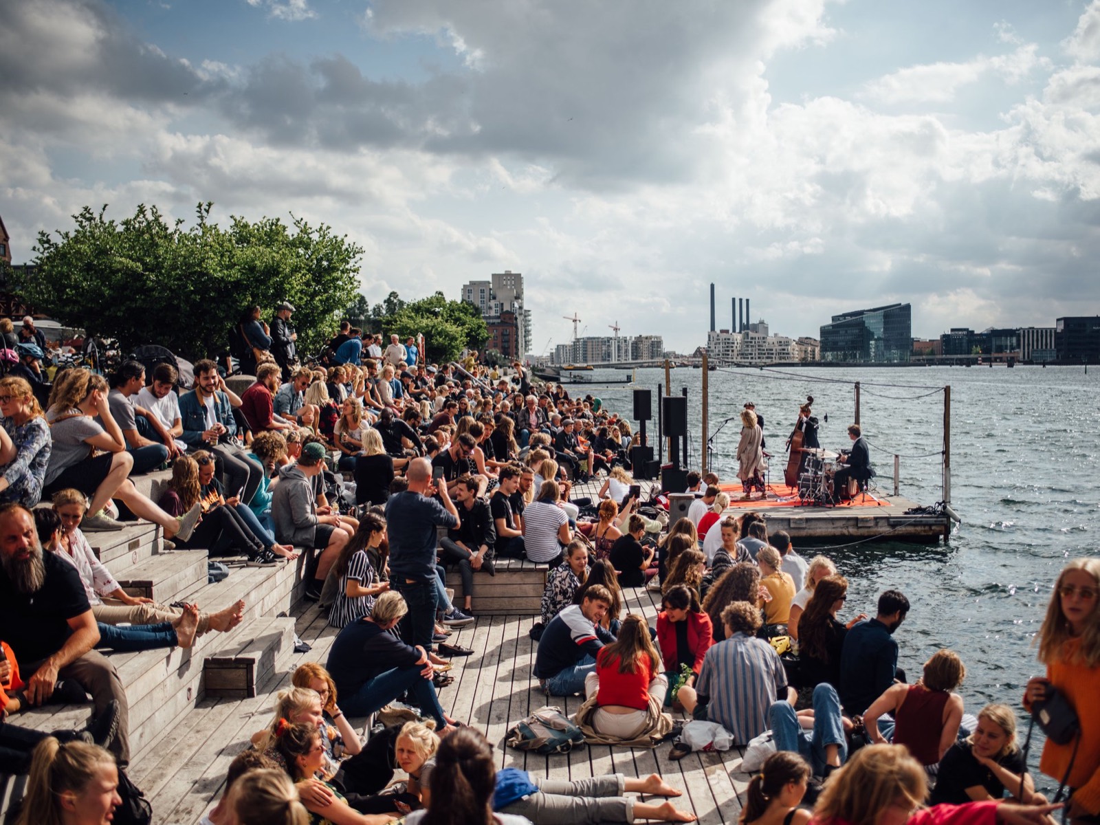 Et stort publikum på havnetrappen lytter til en koncert på en tømmerflåde