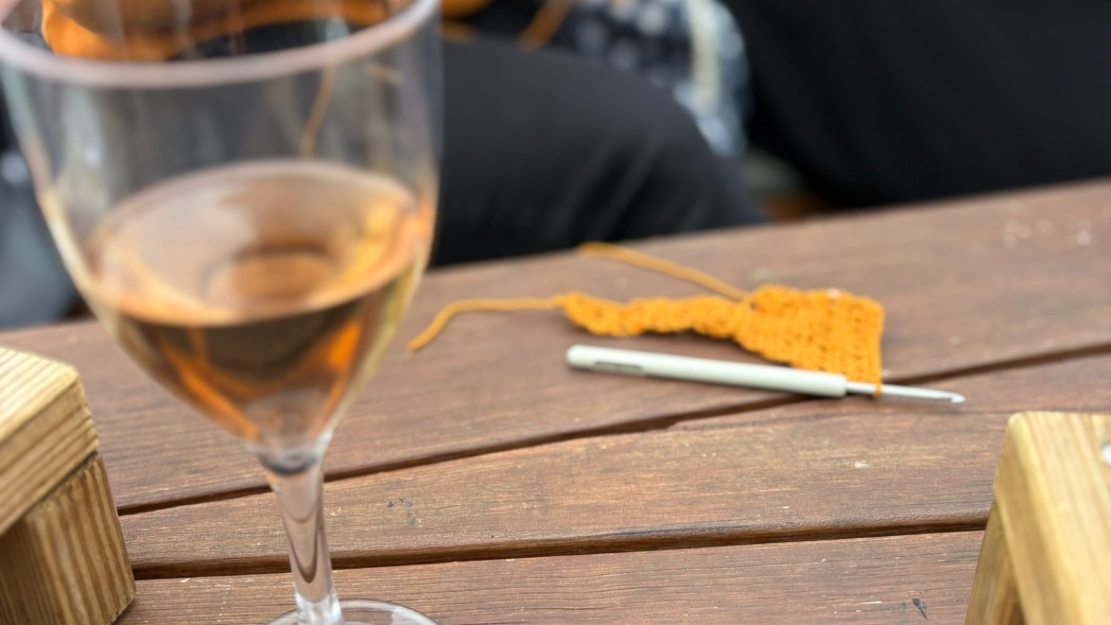 Et glas rosé ved siden af et hæklet stykke og en hæklenål på bordet om bord